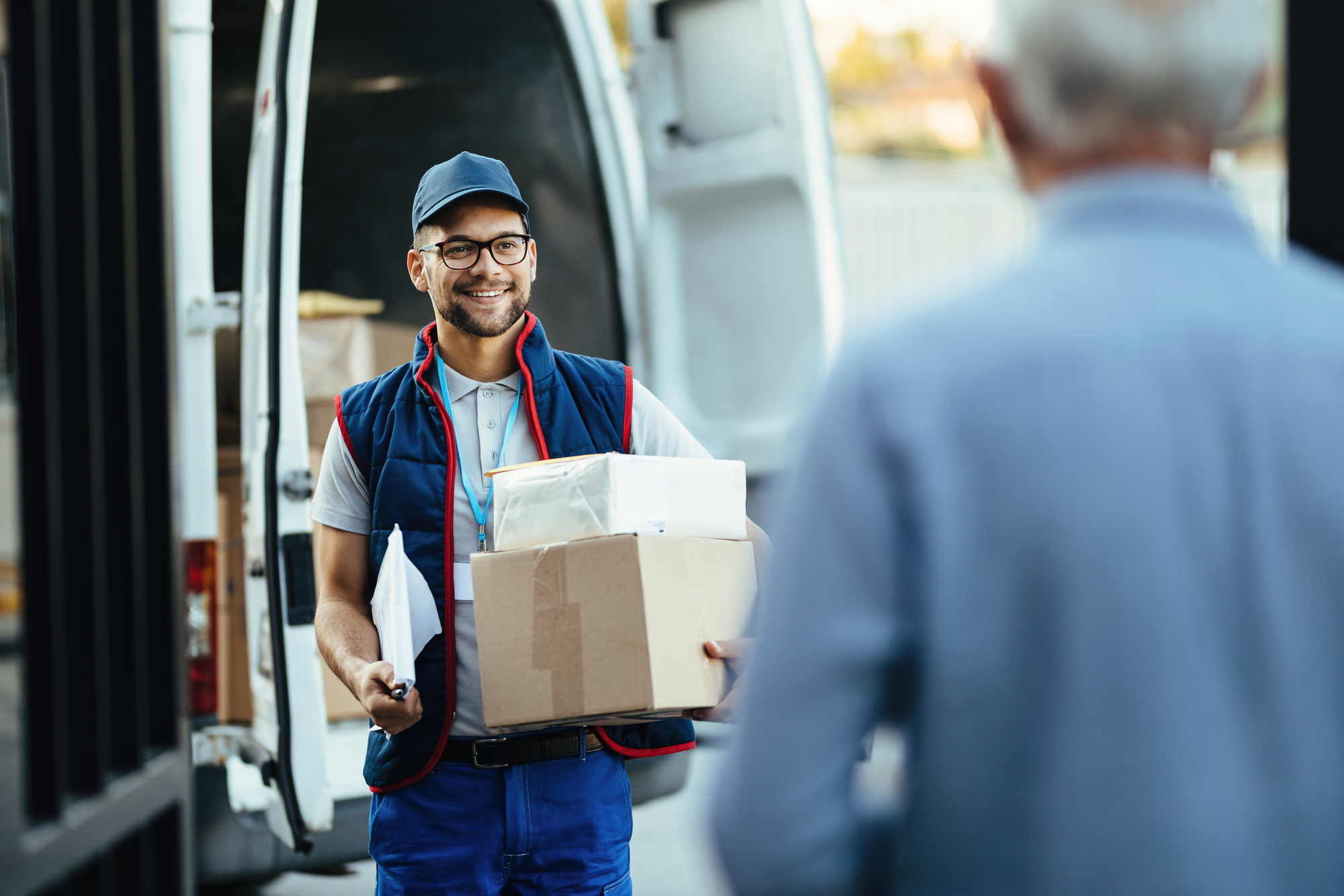 A smiling man stands beside a delivery van, holding a box, ready to ma